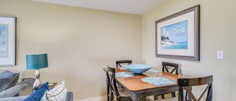 A dining area with a wooden table, chairs, and a bowl. There is a couch with cushions and a lamp on a side table.