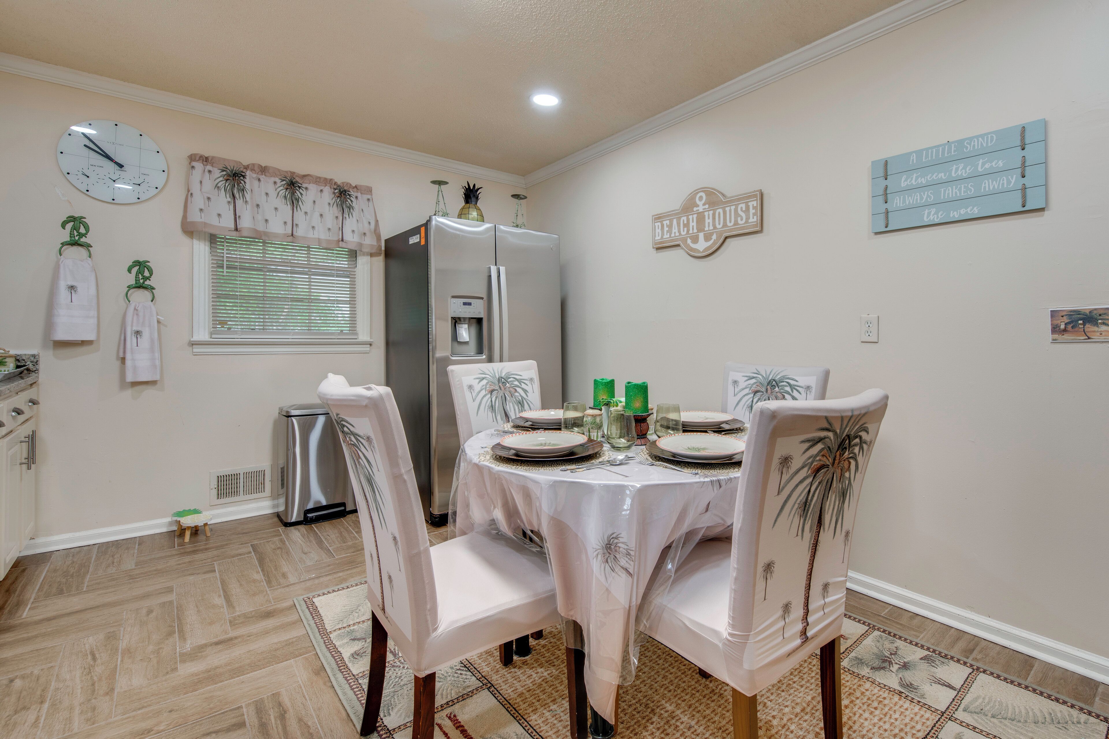 Un comedor con una mesa dispuesta para cuatro, sillas con diseños de palmeras, un refrigerador, un reloj y un cartel en la pared que dice 'BEACH HOUSE'.