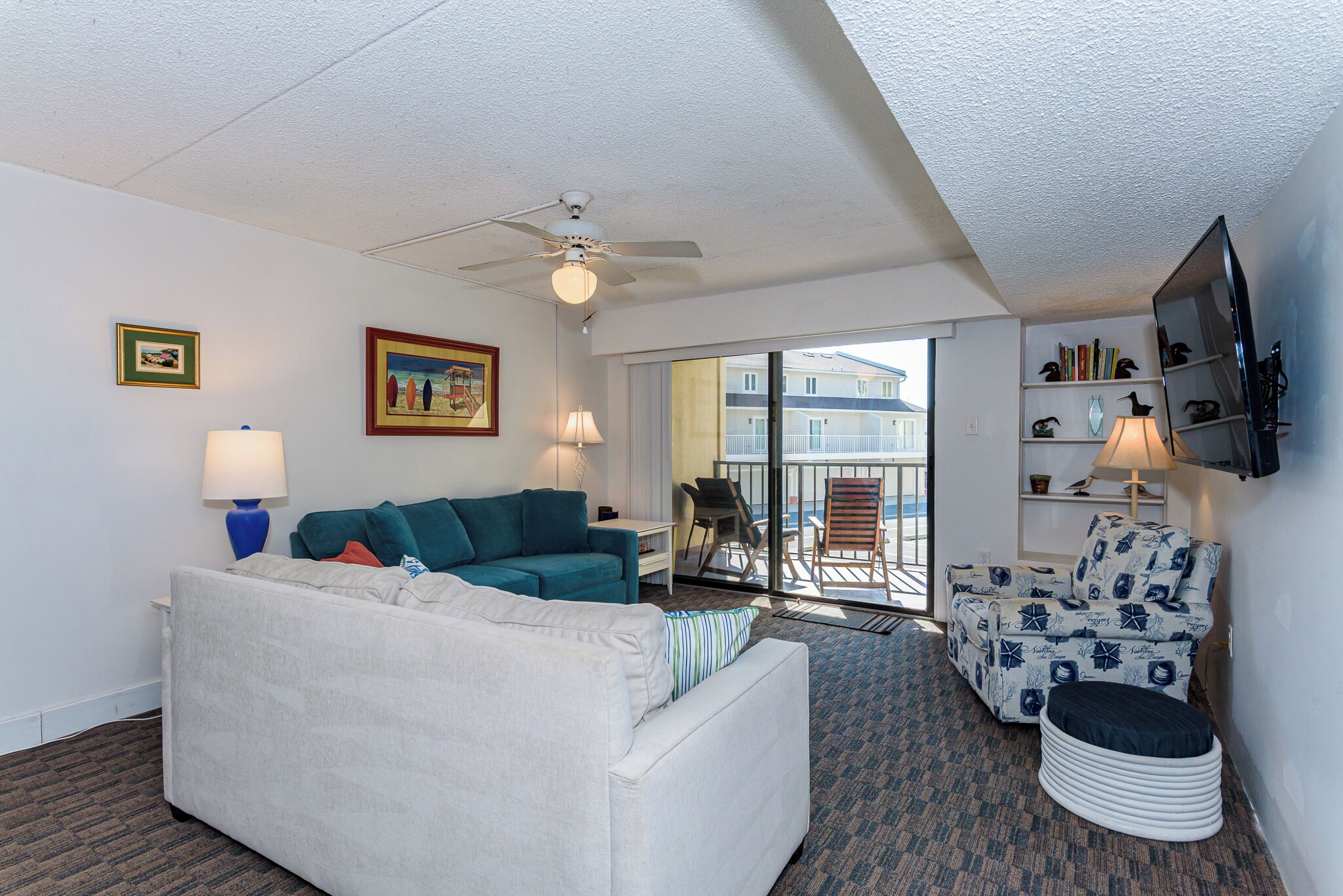 Habitación de hotel con sofá blanco, sillón turquesa y reposapiés estampado. Cuenta con ventilador de techo, televisor montado en la pared y balcón con vistas a edificios.