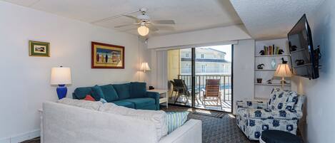 Habitación de hotel con sofá blanco, sillón turquesa y reposapiés estampado. Cuenta con ventilador de techo, televisor montado en la pared y balcón con vistas a edificios.