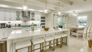 A modern kitchen with white cabinetry, a large island with bar stools, pendant lighting, and a dining area with white chairs. - Luxe Canal Home 2 King Suites Huge Salt Pool (Punta Gorda)
