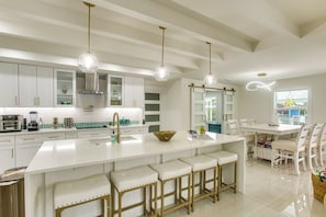 A modern kitchen with white cabinetry, a large island with bar stools, pendant lighting, and a dining area with white chairs. - Luxe Canal Home 2 King Suites Huge Salt Pool (Punta Gorda)