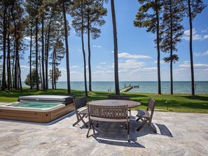 A patio with a hot tub, wooden table, and chairs, overlooking a body of water with tall trees and a dock. - Bayview 2.0 | an ESVR property (Wittman)
