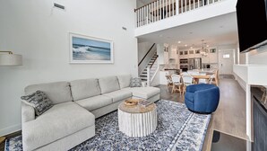 Ett rymligt vardagsrum med en grå soffa, ett runt soffbord och en blå stol. En trappa leder upp till ett köksområde med vita skåp och ett matbord.