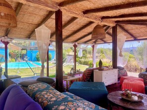 A covered outdoor seating area with a pool view, a dining table with a tray, and a bed with a red bedspread.