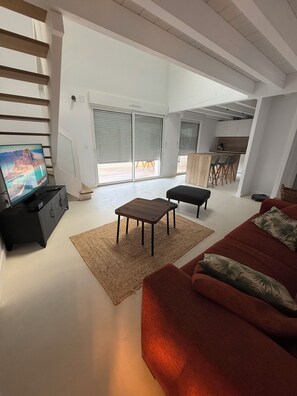 A modern living room with a flat-screen TV, a red sofa, a wooden coffee table, and a kitchen in the background.