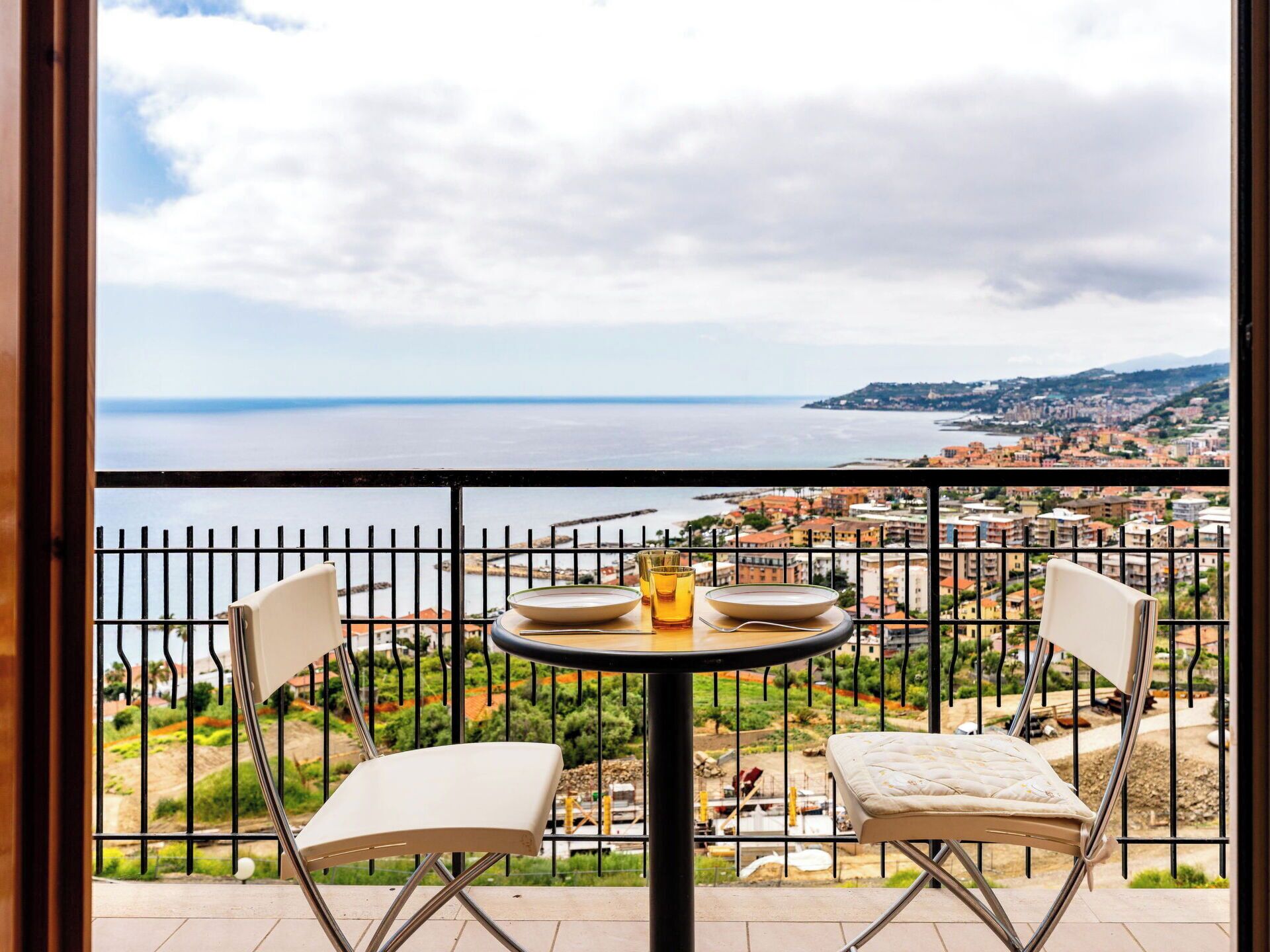 A balcony with a table and chairs, overlooking a coastal cityscape.