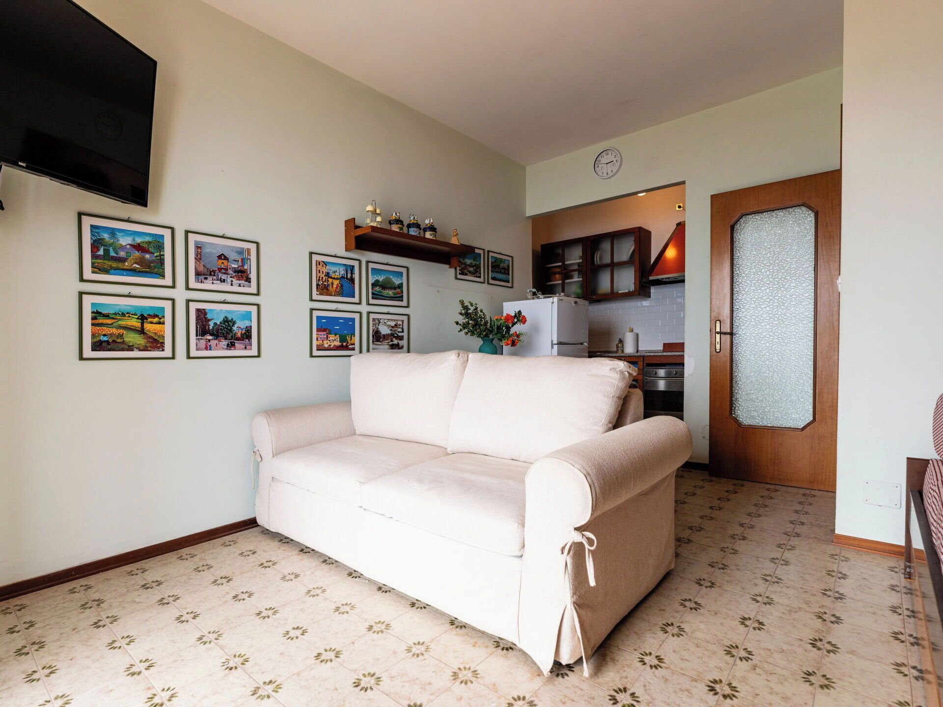 A living room with a sofa, a flat-screen TV, and a kitchen in the background.