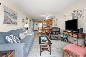 Ein Wohnzimmer mit Sofa, Sessel, Couchtisch, Fernseher und einer Küche im Hintergrund.