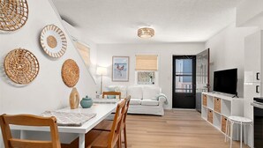 Uma sala de estar moderna com área de jantar, móveis brancos, televisão e objetos decorativos de tecido na parede.