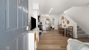 Cozinha moderna com televisão, armários brancos e sala de jantar com cadeiras e mesa de madeira.