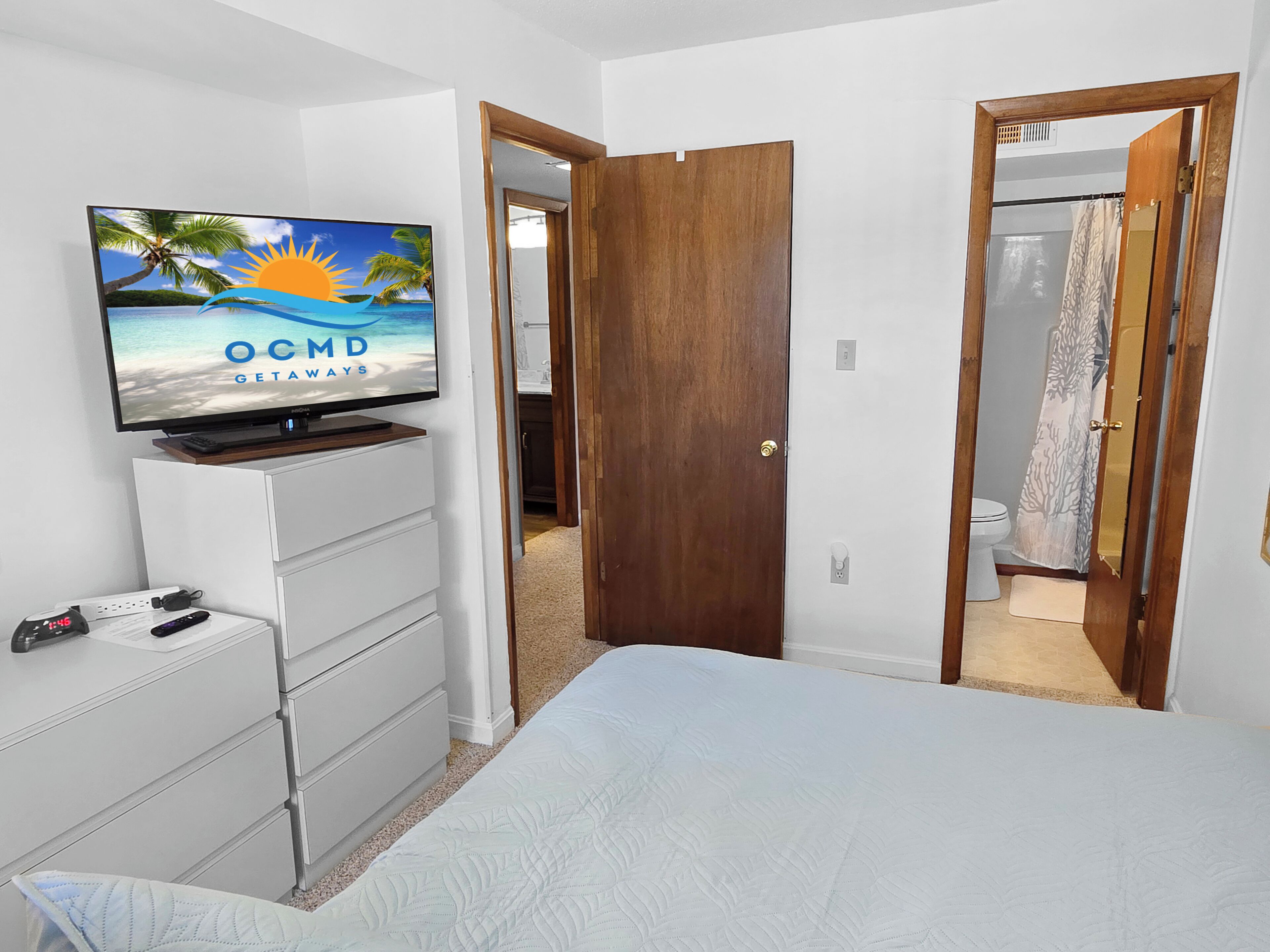 Ein Schlafzimmer mit einem Bett, einem Fernseher auf einer Kommode, einer Holztür und einem angrenzenden Badezimmer.