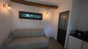 A small, cozy room with a sofa, a window with blinds, a door, and a kitchenette area.