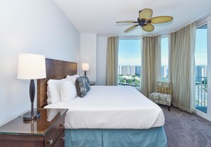 Ein Hotelzimmer mit einem großen Bett, einem Deckenventilator, einem Nachttisch mit Lampe, einem Sessel und Blick auf die Stadt durch große Fenster.
