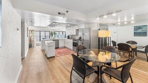 A modern kitchen with white cabinets, a stainless steel refrigerator, and a dining area with wicker chairs and a glass table. - Plaza 1612 2bd Oceanfront condo (Ocean City)