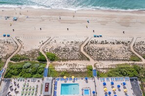 Luftaufnahme eines Strandes mit Poolbereich, inklusive Liegestühlen und Sonnenschirmen.