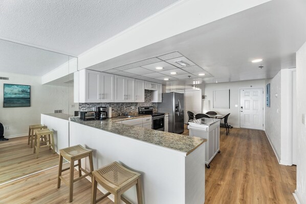 A modern kitchen with white cabinets, granite countertops, and stainless steel appliances. There are wooden stools and a small island with a sink. - Plaza 1612 2bd Oceanfront condo (Ocean City)