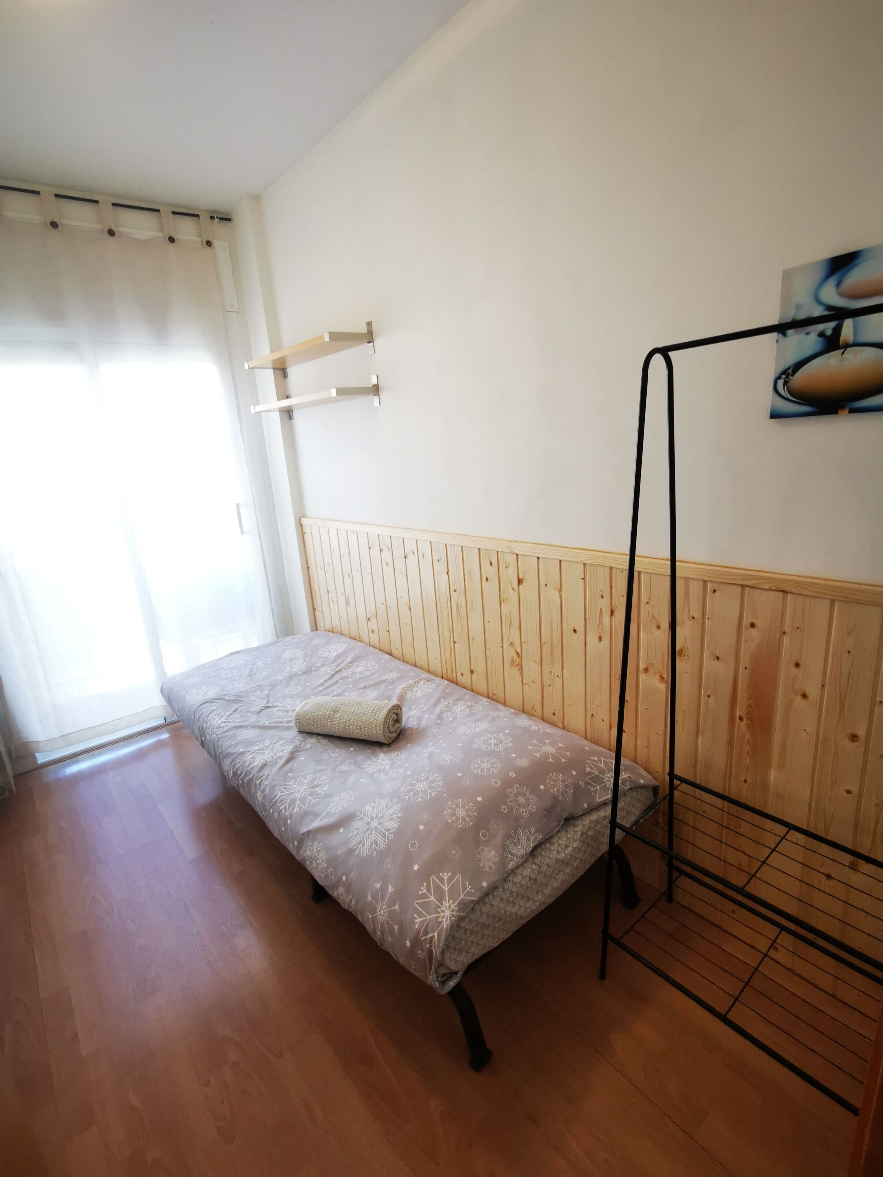A single bed with a patterned bedspread, a wooden headboard, a metal stand, and a wall-mounted shelf.