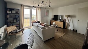 Olohuoneessa on sohva, ruokapöytä tuoleineen, televisio, kirjahylly ja parveke, jossa on verhot.