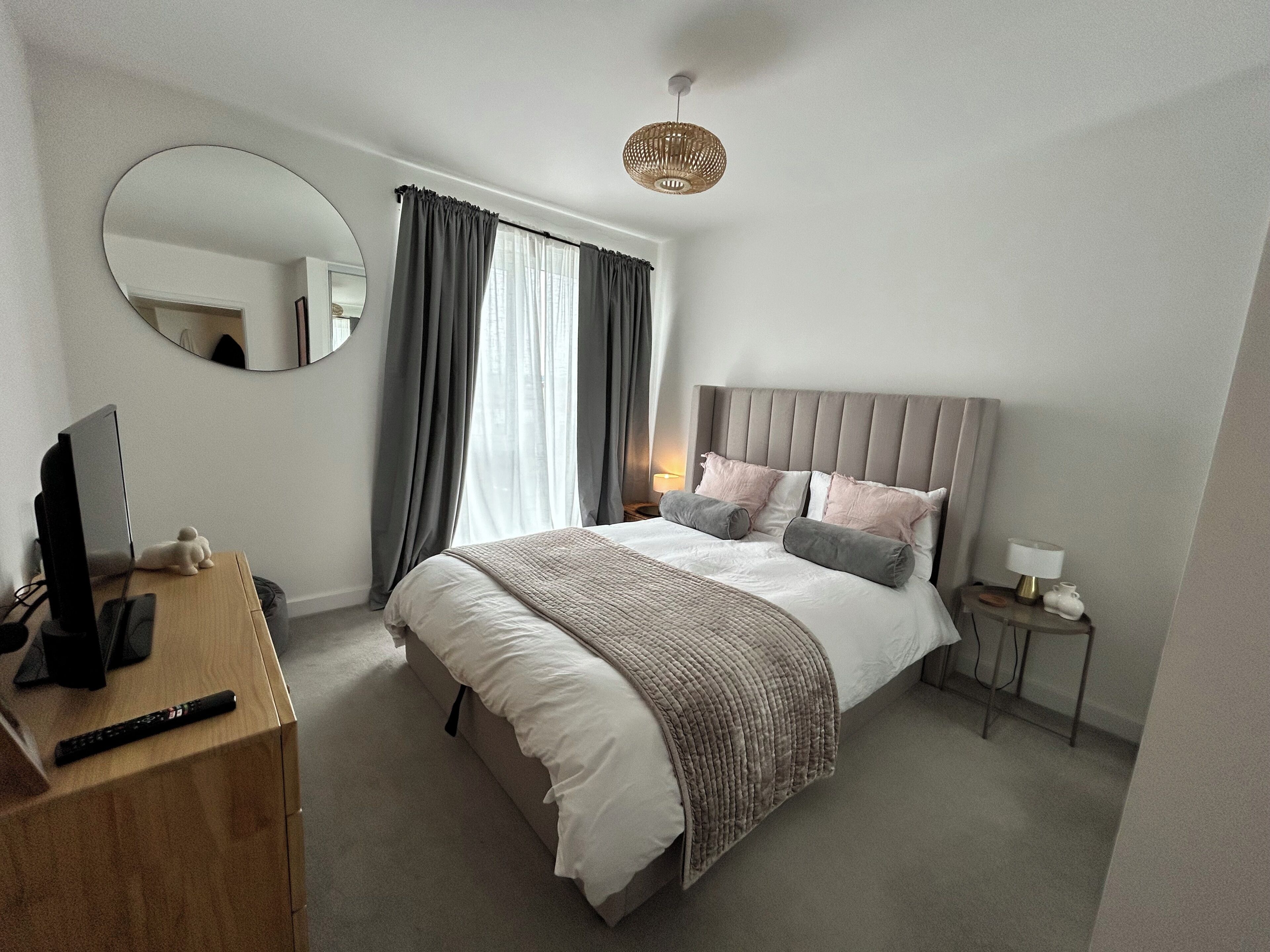 Ein Hotelzimmer mit einem Bett, einem Schreibtisch mit Computer, einem Spiegel, einem Nachttisch mit Lampe und einem Fenster mit Vorhängen.