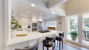 A modern kitchen with white cabinetry, stainless steel appliances, a large island with bar stools, and a view of greenery outside. - 15 Rum Row: 3  BR, 3  BA House in Hilton Head Island, Sleeps 6 (Hilton Head Island)