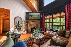 A cozy living room with a fireplace, leather sofa, and a large window with a view of greenery. - Creekfront 4BR Cabin: Hot Tub, Sauna, Dome, Game Room & Slide (Sevierville)