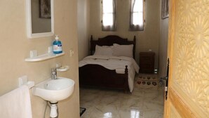 A bedroom with a bed, a bedside table, a nightstand, a mirror, a shelf with toiletries, and a window with curtains. - Dar Margoum (Chwiter)