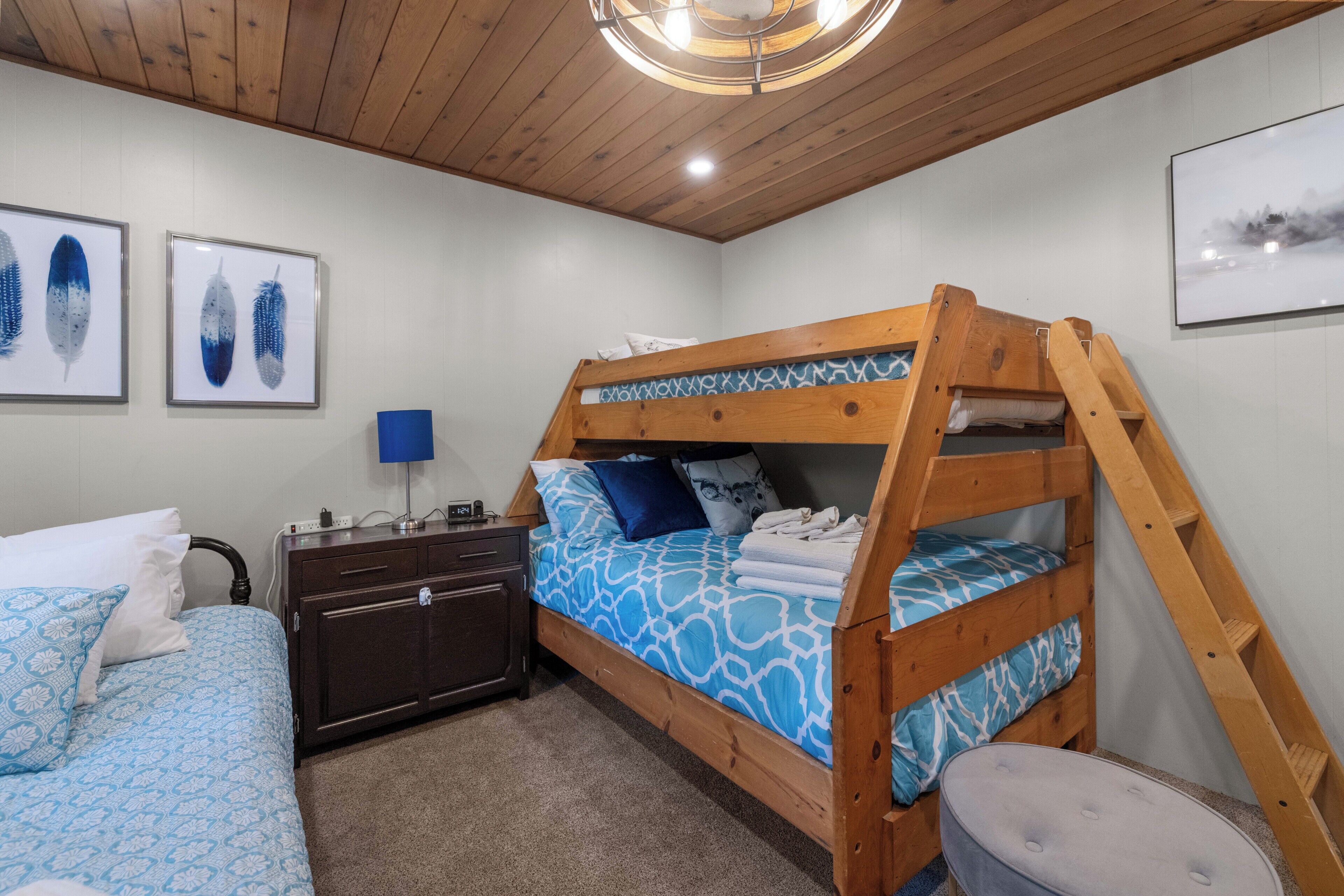 Een stapelbed met een blauw bedlaken met patroon, een houten dressoir, een bureau met lamp en lijstjes met kunstwerken aan de muur.