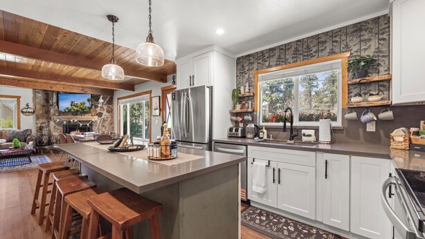 A modern kitchen with a large island, wooden bar stools, a stone fireplace, and a window with a view of the outdoors. - Cozy Bear Lodge (Big Bear City)