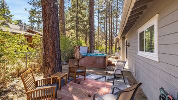 A cabin with a hot tub, wooden chairs, and a patio area surrounded by tall trees.
