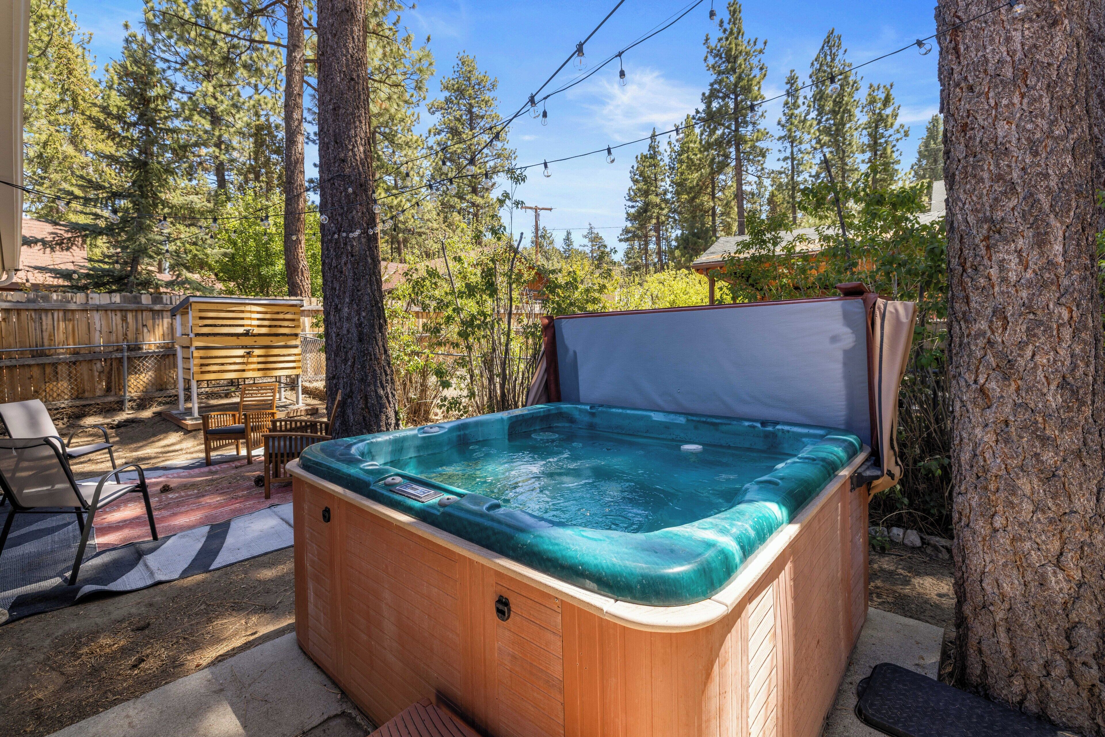 A hot tub in a backyard with a wooden deck, chairs, and a tree.