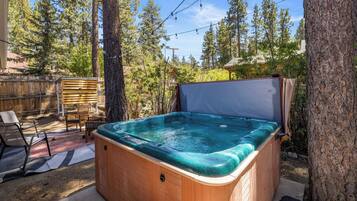 A hot tub in a backyard with a wooden deck, chairs, and a tree.