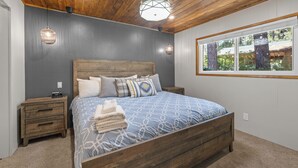 A bedroom with a wooden ceiling, a large bed with patterned bedding, two bedside tables, and a window with a view of trees. - Cozy Bear Lodge (Big Bear City)