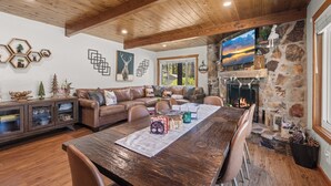 A cozy living room with a wooden ceiling, a stone fireplace, a large TV, and a dining table with chairs. - Cozy Bear Lodge (Big Bear City)