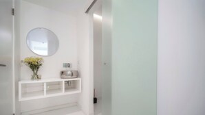 Una pared blanca con un espejo redondo, un pequeño estante con un florero y una tetera.