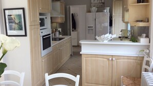Una cocina con armarios de madera, encimera blanca, microondas, horno, refrigerador y una zona de comedor con una silla y una mesa.