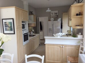 A kitchen with wooden cabinets, a white countertop, a microwave, an oven, a refrigerator, and a dining area with a chair and a table. - Royan: Bright family villa for 11 people - 7 min from Pontaillac beach (Royan)