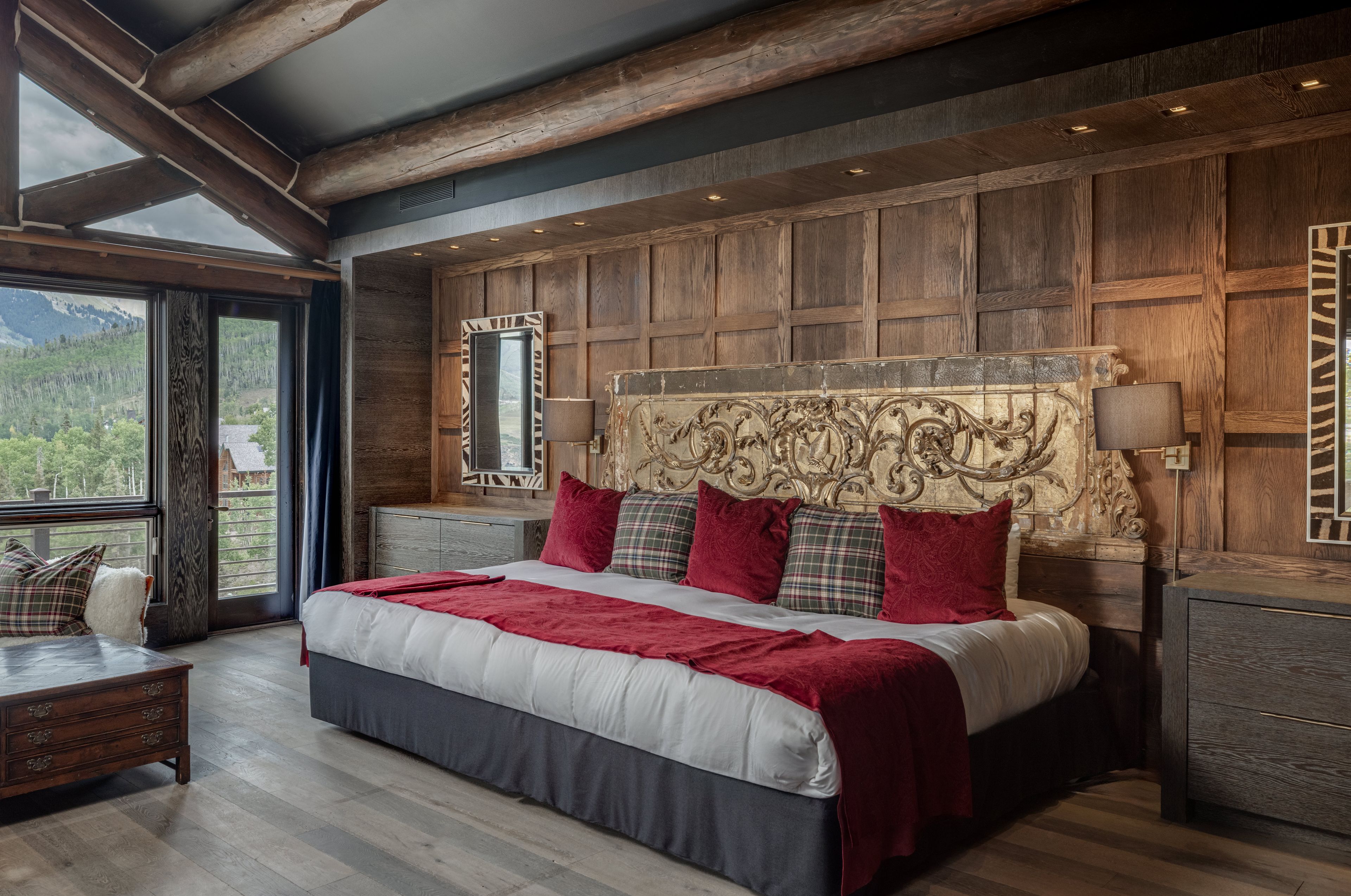 A large bed with a wooden headboard, a nightstand, and a view of a mountainous landscape through a large window.
