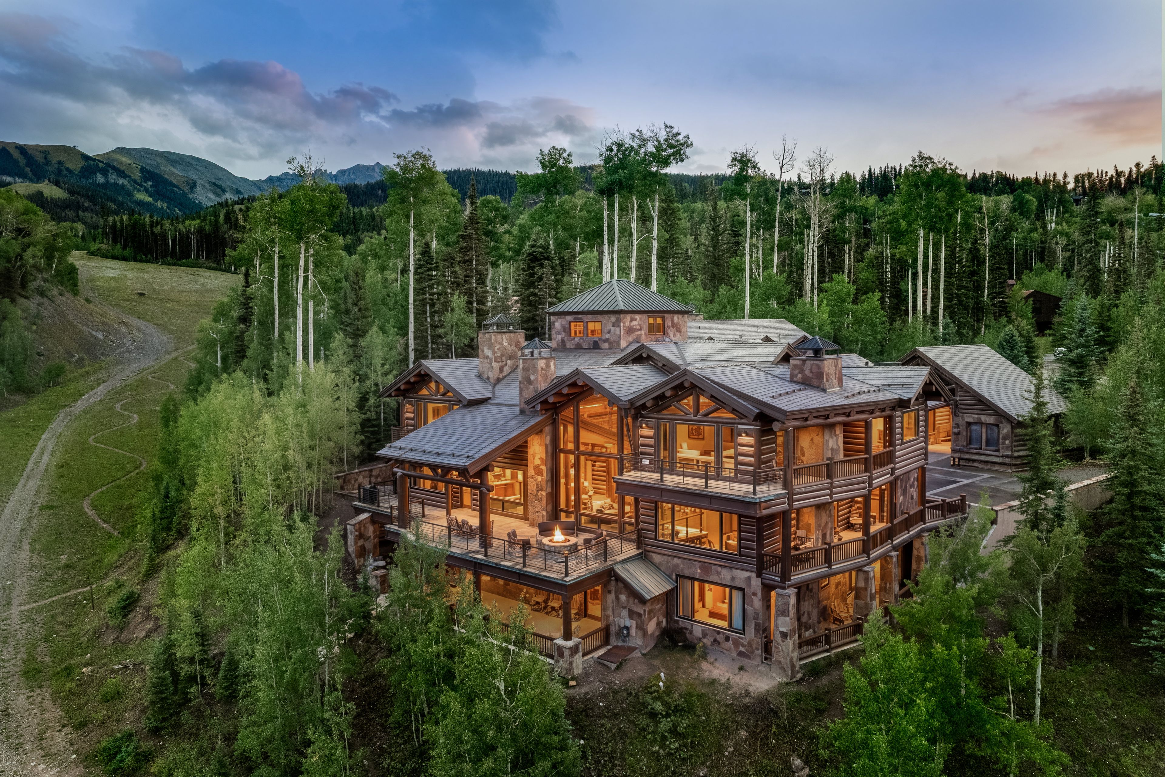 A large log cabin with multiple levels and a fireplace, surrounded by a forest and mountains.