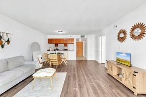 Ein modernes Wohnzimmer mit einem weißen Sofa, einem Essbereich mit Holzsesseln, einer Küche mit Holzschränken und einem Fernseher auf einem Holzschrank.