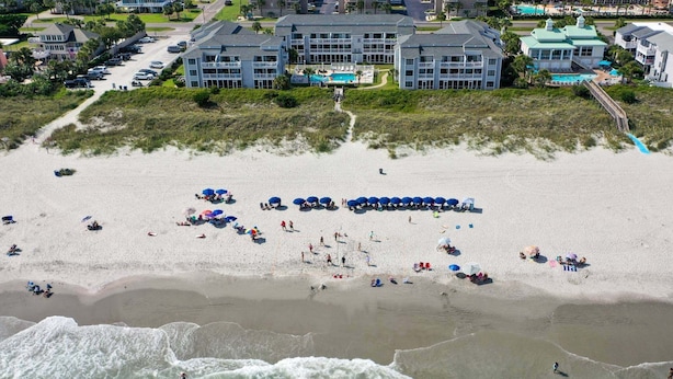 Ein Strandhotel mit mehreren Gebäuden, einem Sandstrand mit Sonnenschirmen und brechenden Wellen.