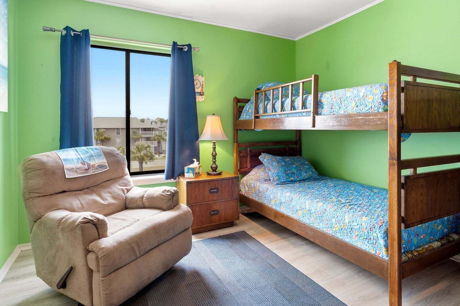 A room with a bunk bed, a chair, a lamp, and a view of the outside.