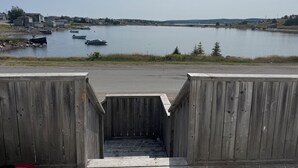 Une jetée en bois mène à un lac paisible où se trouvent des bateaux, le tout étant entouré de maisons et de collines.
