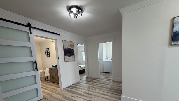 Un couloir avec une porte coulissante, un luminaire au plafond et une vue sur une chambre à coucher où l’on aperçoit un lit et un lave-linge.