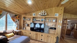 A wooden cabin interior with a TV, shelves with books and decorations, a sofa, and a window with curtains. - Queens Way Retreat-12 Guests-Pet Friendly (Wells)