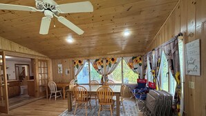 A wooden cabin interior with a dining area, ceiling fan, and windows with sunflowers. - Queens Way Retreat-12 Guests-Pet Friendly (Wells)