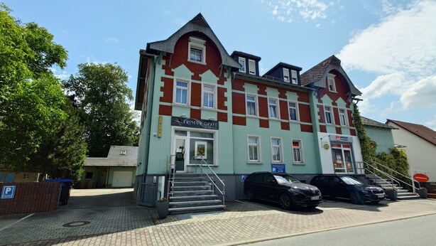 A multi-story building with a sign reading "Friseur Graft" on the ground floor, indicating a hair salon. There are steps leading to the entrance, and two cars parked in front.