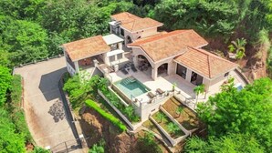 A large, multi-story house with a swimming pool, surrounded by lush greenery.