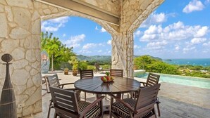 A stone-arched outdoor dining area with a round table and chairs, overlooking a pool and scenic landscape.