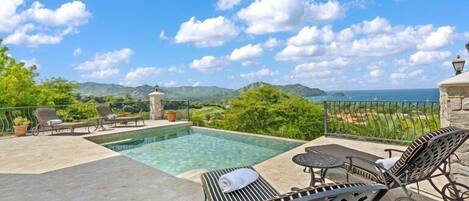 A pool area with lounge chairs and a table, overlooking a mountainous landscape and the sea.